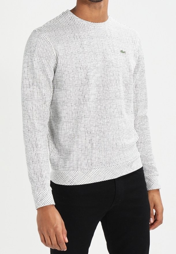 Homme portant une chemise blanche à manches longues avec un petit motif à grille noire et un pantalon noir sur fond uni.
