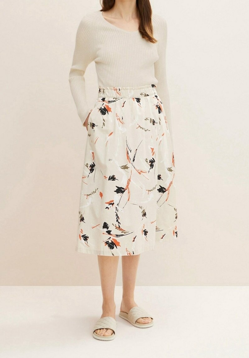 Femme portant un haut blanc à manches longues côtelé et une jupe midi blanche avec des motifs floraux noirs, orange et olive, debout, les mains dans les poches.