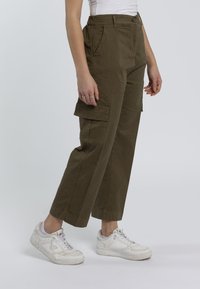 Pantaloni cargo khaki con vita alta, tasche laterali e gambe larghe. Indossati con scarpe da ginnastica bianche su uno sfondo neutro.