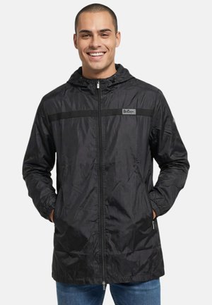 Homme souriant portant une veste à capuche noire zippée avec des poches latérales et un jean bleu, debout les mains dans les poches.