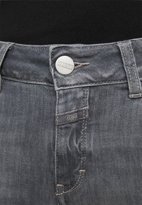 Szare dżinsy z denimu z zapięciem na srebrny guzik, widocznymi detalami szycia i metką "CLOSED" przy pasku. Gładka tekstura.