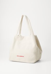 Bēšs audekla iepirkumu soma ar divām rokturiem, kurai ir izteikts sarkans "KARL LAGERFELD" logo apakšējā daļā. Strukturēts, plašs dizains.