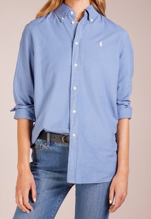 Personne portant une chemise bleu clair à manches longues avec les poignets retroussés, un jean bleu et une ceinture noire, cadrée au niveau du cou et des genoux.