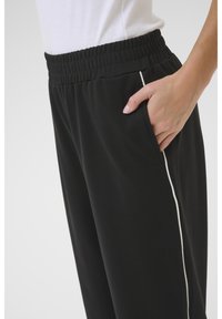 Pantaloni da tuta neri con cintura elastica e tasca laterale, con una rifinitura bianca lungo la cucitura esterna. Texture del tessuto liscia.