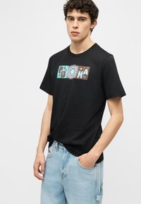 Νέος άνδρας με σγουρά μαλλιά που φοράει μαύρο T-shirt με πολύχρωμο λογότυπο "PUMA" με λουλούδια και ανοιχτόχρωμο τζιν μπλούζα, σε απλό φόντο.