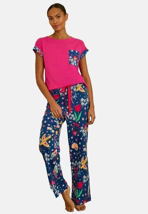 Femeie purtând un tricou roz aprins cu mânecă scurtă cu buzunar floral și pantaloni de pijama albaștri cu modele florale și stelate colorate, stând desculță.