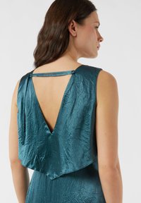 Turkusowa sukienka z głębokim dekoltem w kształcie litery V na plecach, wykonana z marszczonej tkaniny. Posiada nałożoną zwiewną warstwę materiału i wąskie ramiączka zapinane na ramionach.