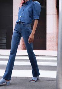 Camisa de denim con mangas abullonadas, jeans acampanados de color azul claro y sandalias de tacón abierto en azul. Tejido suave, diseño casual y corte ajustado.