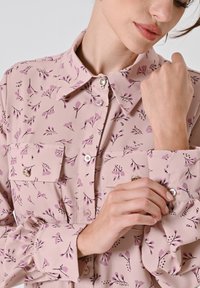Blusa rosa con stampa floreale, caratterizzata da due tasche sul petto, colletto con bottoni e polsini arricciati. Dettagli in tessuto liscio e leggero.