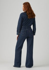 Une salopette en denim bleu foncé avec des manches longues, une taille ajustée, des pantalons à jambes larges, et deux poches arrière avec des détails de couture visibles.