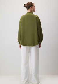 Olive zielona oversize kurtka z wysokim kołnierzem i długimi rękawami, w zestawieniu z białymi spodniami o szerokich nogawkach. Gładka struktura, casualowy design.