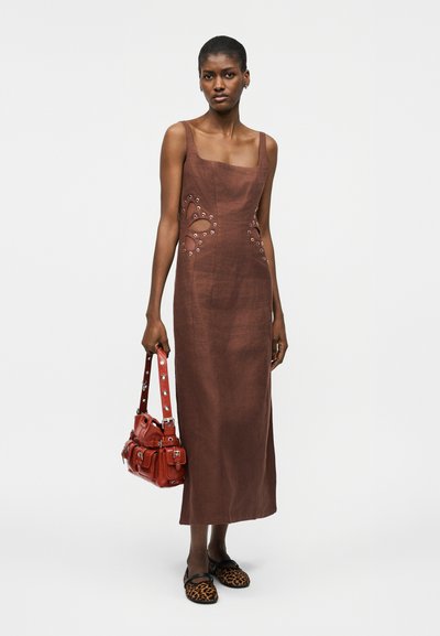 Γυναίκα που φοράει καφέ αμάνικο φόρεμα με κοψίματα, κρατώντας μια κόκκινη τσάντα, σε συνδυασμό με μοκασίνια με λεοπάρ μοτίβο, στέκεται μπροστά από λευκό φόντο.