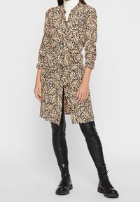 Abito a fantasia floreale in nero con fiori gialli e rosa; presenta un colletto, bottoni e una cintura da annodare, indossato sopra pantaloni neri aderenti.