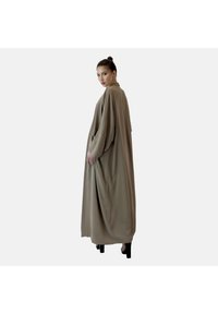 Longue cape beige fluide avec de larges manches, un col haut et des fentes latérales, fabriquée dans un tissu lisse. Le modèle se tient de profil contre un fond clair.