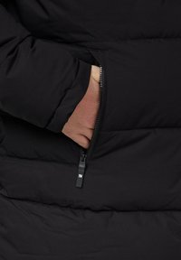 Schwarze gepolsterte Jacke mit einer sichtbaren Reißverschluss-Tasche und einer Hand, die hineingesteckt ist. Der Stoff wirkt glatt, hat eine weiche Textur und isolierte Schichten.