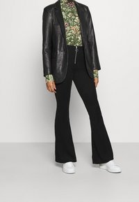 Svart skinnblazer över en grön mönstrad polotröja, tillsammans med utsvängda svarta ribbade byxor och vita sneakers. Enkel dragkedjedetalj syns.