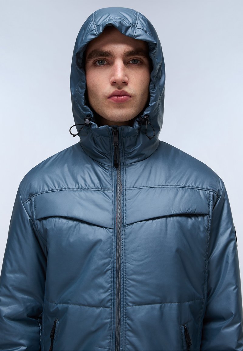 Chaqueta acolchada azul con capucha, que presenta un cierre de cremallera y cordones elásticos. El material tiene un acabado suave y brillante con textura acolchada.