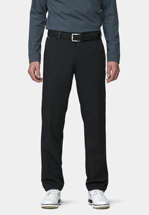 Mann trägt schwarze gerade geschnittene Hose, schwarzen gewebten Gürtel, dunkelgraues Langarmshirt und weiße Golfschuhe, steht vor einfarbigem Hintergrund.