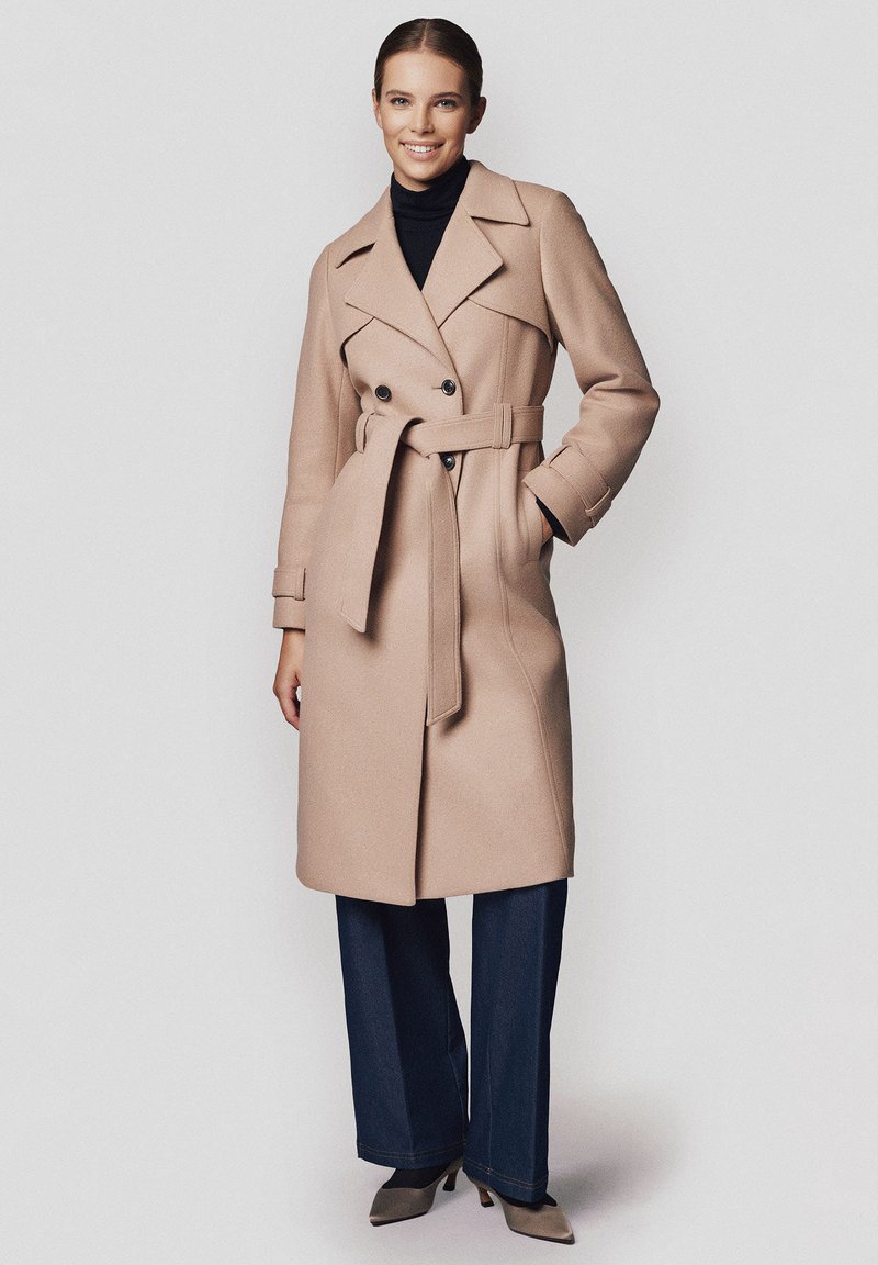 Beżowy wełniany płaszcz o podwójnym zapięciu, z szerokimi klapami i paskiem w talii. Połączony z ciemnymi spodniami o szerokich nogawkach i spiczastymi butami.