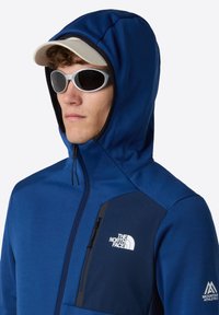 Blauwe hoodie gemaakt van zachte stof met een ritszak, voorzien van een logo op de borst en contrasterende zwarte accenten.