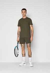 Olijfgroen sport T-shirt en korte broek, van katoen, met korte mouwen en een gebandeerde halslijn. Witte sportschoenen, met een tennisracket in de hand.