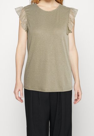 Femme portant un haut sans manches vert olive avec des manches en dentelle transparente à volants et un pantalon noir ample, debout devant un fond uni.