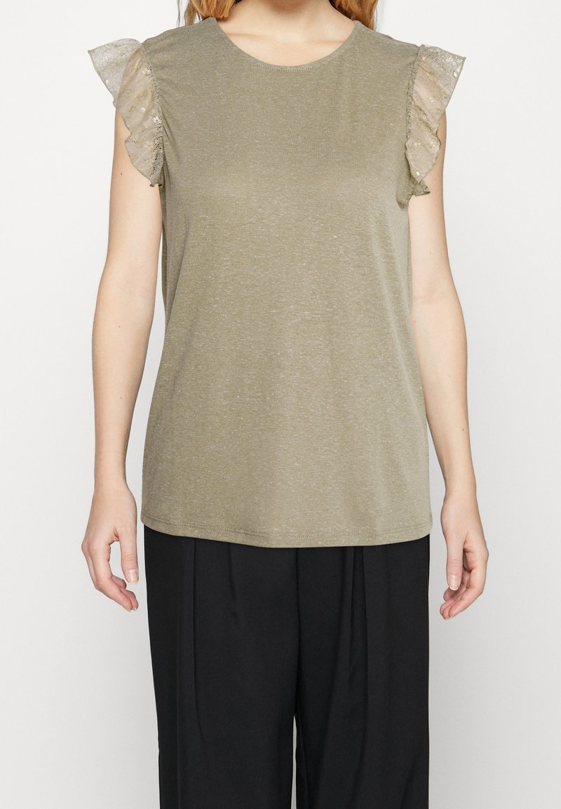Femme portant un haut sans manches vert olive avec des manches en dentelle transparente à volants et un pantalon noir ample, debout devant un fond uni.