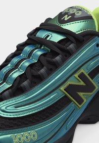 Κοντινό πλάνο ενός μαύρου και ιριδίζοντος μπλε-πράσινου αθλητικού παπουτσιού με μαύρα κορδόνια, κίτρινο-πράσινο λογότυπο "N" και λεπτομέρεια "1000" κοντά στη σόλα.