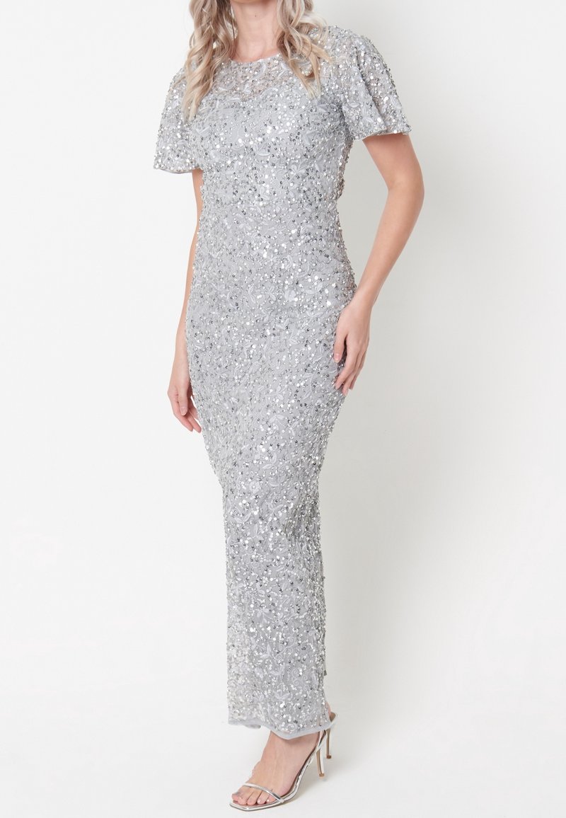 Femme portant une robe argentée longue au sol à manches courtes en sequins avec des sandales argentées à talons hauts, debout sur un fond uni.