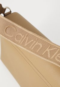 Sac bandoulière beige avec une surface texturée, doté d'une large sangle embossée du nom "Calvin Klein" dans une couleur assortie.