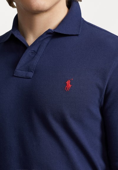 Granatowa koszulka polo z fakturowanej tkaniny, zapinana na trzy guziki, z czerwonym haftowanym logo po lewej stronie na piersi. Krótkie rękawy.