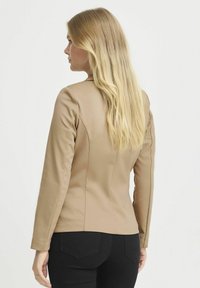 Blazer aderente beige con maniche lunghe, tessuto liscio, cuciture verticali e spalle strutturate. Abbinato a pantaloni neri.