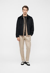 Veste bomber en daim noir portée sur une chemise à boutons beige, associée à un pantalon beige clair et des chaussures noires et blanches.