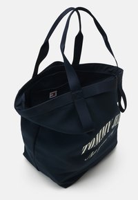 Sac fourre-tout bleu marine Tommy Jeans avec doubles poignées et texte de logo blanc sur le côté, présenté ouvert et vide sur fond blanc.