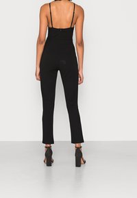 Kvinna iförd en svart ärmlös jumpsuit med dragkedja i ryggen och svarta högklackade sandaler, stående mot en enkel ljus bakgrund.