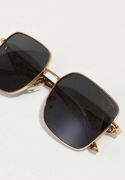 Gafas de sol cuadradas con lentes oscuros, montura de metal dorado y un intrincado diseño calado en las patillas. Presenta acentos angulados en las varillas.
