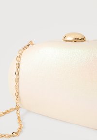 Πέρλα λευκό οβάλ τσαντάκι χειρός με λαμπερό χρυσό κούμπωμα και ταιριαστή χρυσή αλυσίδα λουρί σε λευκό φόντο.