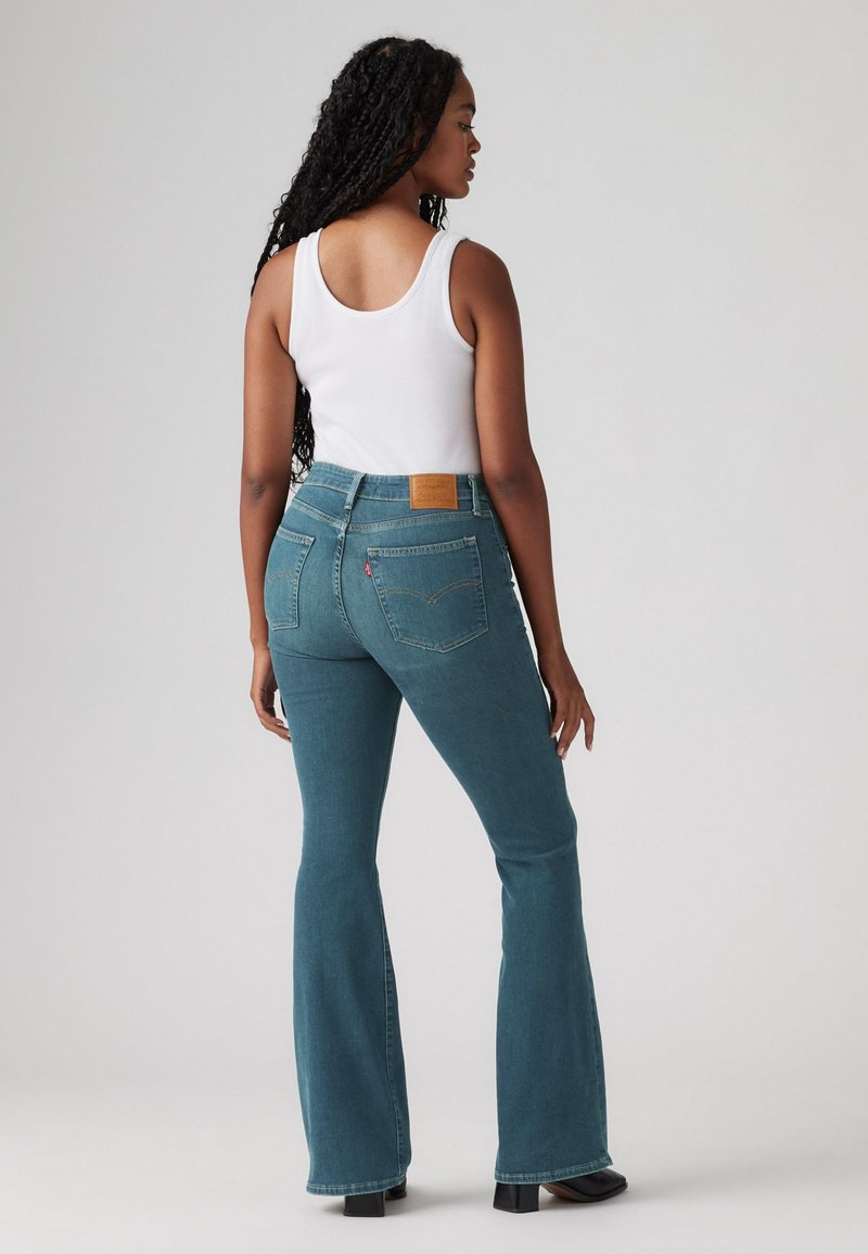 Jeans a zampa d'elefante in denim teal con vita alta e tasche posteriori a toppa. Il modello indossa una canotta bianca e stivaletti neri.