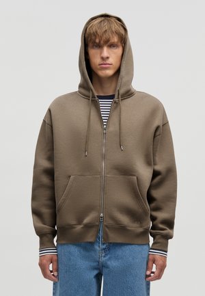 Młody mężczyzna w brązowej bluzie na zamek z troczkami, nałożonej na pasiastą koszulę i niebieskie dżinsy, stojący na jednolitym tle.