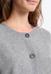 Femme portant un cardigan tricoté gris avec de grands boutons ronds noirs, montrant son cou, son menton et une partie de ses lèvres.