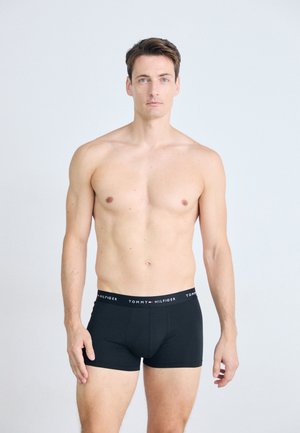 Oberkörperfreier Mann mit kurzen braunen Haaren, der schwarze Tommy Hilfiger Boxershorts trägt und vor einem schlichten, hellgrauen Hintergrund steht.