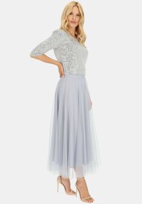 Srebrna bluzka z cekinami i rękawami do trzy czwarte, połączona z jasnoniebieską, zwiewną spódnicą z tiulu. Modelka nosi beżowe sandały na obcasie.