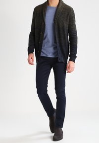 Tmavě zelený kardigan s šálovým límcem a knoflíky, přes kterým je lehké modré tričko. Námořnické slim-fit kalhoty a tmavě šedé boty dotvářejí celkový vzhled.