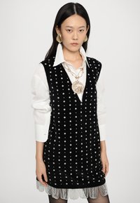 Μαύρο βελούδινο φόρεμα διακοσμημένο με λευκά λουλουδάτα μοτίβα, με v-λαιμόκοψη, στρωμένο πάνω από λευκό πουκάμισο, ολοκληρωμένο με περιθώριο με φράντζα. Χρυσά κοσμήματα ως αξεσουάρ.