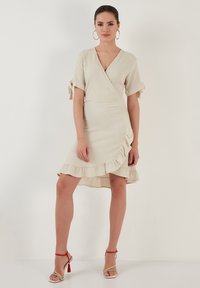 Light beige wrap dress with short sleeves, tied accents, and ruffled hem. Worn with multicolored heeled sandals. Smooth texture.
