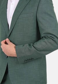 Blazer verde con texture, dotato di una tasca sul petto e polsini abbottonati. Il tessuto presenta un motivo sottile e una finitura liscia.