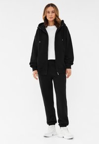 Zwarte rits hoodie met koorden, gedragen over een wit T-shirt, gecombineerd met zwarte joggingbroeken en witte sneakers. Glad materiaal, relaxte pasvorm.