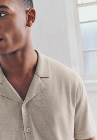 Beige Polohemd mit kurzen Ärmeln aus strukturiertem Stoff, mit einem abgeschnittenen Kragen und zwei Knöpfen. Der Hintergrund ist eine hellfarbige Wand.