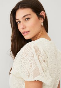 Hvid, blonderet bluse med puffærmer, der har indviklede blomster mønstre og et tekstureret design. Glat stof, beskeden udskæring og elegante detaljer.