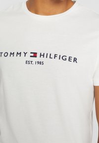 Bílý bavlněný tričko s výšivkou loga "TOMMY HILFIGER" v námořnické modré a červené barvě, pod kterým je nápis "EST. 1985"; kulatý výstřih a krátké rukávy.